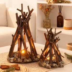 Op de afbeelding: Twee decoratieve kandelaars in de vorm van miniatuurkampvuren. Elk heeft een basis van grijze stenen die een tipi van bruine houten takken ondersteunt. Een warm, geel licht straalt van binnenuit, waardoor een gezellige sfeer ontstaat. De kandelaars staan op een neutrale ondergrond.