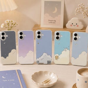 Peut inclure: Cinq coques de téléphone avec des motifs de nuages et de lune dans différentes couleurs : gris foncé, lavande, bleu clair et bleu ciel. Un étui présente un petit ours. Un carnet avec le texte "Take it easy" est également visible.
