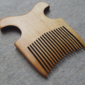 May include: A wooden comb with a handle and a wide-tooth design. The comb is made of light-colored wood with a natural grain pattern. The handle has a curved shape for easy grip. The comb's teeth are evenly spaced and dark in color.