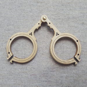 Può includere: Un oggetto in legno chiaro, possibilmente un oggetto decorativo o uno strumento, con due componenti circolari collegati da una cerniera. Ogni parte circolare presenta un design decorativo e una piccola apertura. L'oggetto è in legno chiaro.
