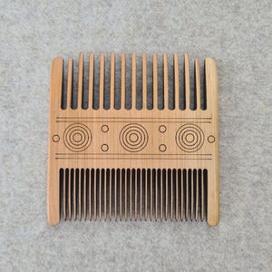 Könnte beinhalten: Ein hellbrauner Holzkamm mit zwei Zahnreihen und einem dekorativen Muster. Das Design umfasst geätzte konzentrische Kreise und kleine Punkte. Der Kamm hat eine rechteckige Form mit abgerundeten Kanten, für die Haarpflege.