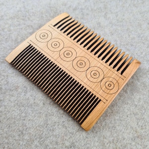 May include: A wooden comb with fine teeth and a decorative design of circles. The comb is made of light-colored wood and has a rectangular shape with rounded edges. The design is etched into the wood.