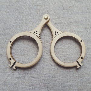 May include: A pair of light-colored wooden eyeglasses with circular frames, connected by a hinge. The frames have decorative cutouts and small holes. The design suggests a vintage or handcrafted aesthetic.