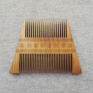 May include: A wooden comb with a trapezoid shape. The comb has two rows of teeth and a decorative band with engraved circles. The wood has a light brown color.