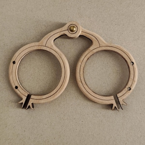 Puede incluir: Una herramienta de madera con dos aberturas circulares conectadas por un mango curvo. Tiene un remache de latón en el punto de conexión y líneas oscuras grabadas en la madera. Las secciones circulares están divididas con un diseño decorativo.