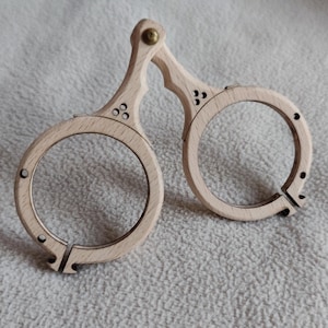 Puede incluir: Un par de gafas de madera de estilo antiguo. Las monturas de madera clara son circulares y están conectadas por una bisagra y un remache de latón. Las monturas tienen agujeros decorativos perforados y un diseño dividido para las lentes.