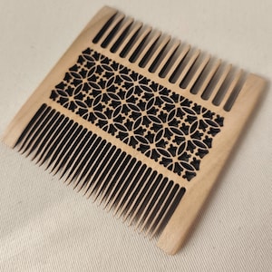 May include: A light brown wooden comb featuring a central rectangular panel with an intricate, laser-cut geometric pattern of interlocking circles and floral shapes. The comb has two rows of fine, evenly spaced teeth on opposite sides.