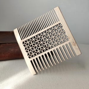 May include: A light-colored wooden comb with a decorative, openwork design. The comb features closely spaced teeth and a central section with a geometric floral pattern. The comb is likely for hair care and grooming.