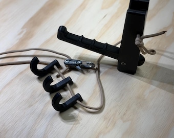 Puede incluir: Equipo negro impreso en 3D con un asa y tres ganchos, conectados por cuerdas color beige y un cierre metálico. El equipo está diseñado para montarse en una superficie, con un asa para agarrar y ganchos para colgar objetos.