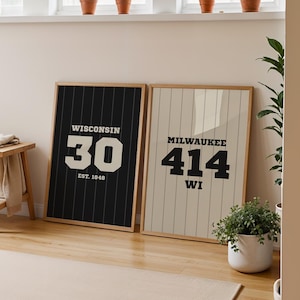 May include: Two framed prints lean against a wall. The left print is black with white text: "WISCONSIN 30 EST. 1940". The right print is cream with black text: "MILWAUKEE 414 WI". The room has a wooden stool, plants, and a rug.