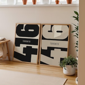 May include: Two framed posters with the number "97" in large black and off-white font, with "TORONTO" below. The posters lean against a wall in a room with a wooden stool, a rug, and potted plants. The room has a neutral color palette.