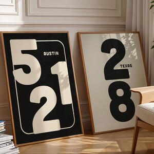 May include: Two framed art prints with large numbers and city names. One print features the number "512" with "Austin" in a black frame. The other print has the number "28" with "Texas" in a black frame. The prints are displayed in a well-lit room.