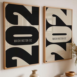 Op de afbeelding: Twee ingelijste kunstprints met een minimalistisch ontwerp. Elke print toont het nummer "202" in zwart en crème, met "WASHINGTON DC" in een schreefloze letter. De lijsten zijn van licht hout. De prints worden boven een houten tafel getoond.