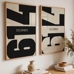 Könnte beinhalten: Zwei rechteckige Wandbilder in hellen Holzrahmen mit einem auffälligen Grafikdesign, das die schwarzen Zahlen 614 und das Wort COLUMBUS in Weiß und Schwarz zeigt. Das Kunstwerk präsentiert eine kontrastreiche Komposition mit großer Typografie.
