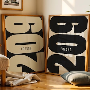 May include: Two framed art prints with the word "FRESNO" in a bold, modern font. The prints feature large black numbers against a cream background. The frames are made of light-coloured wood. A framed picture of palm trees is visible in the background.