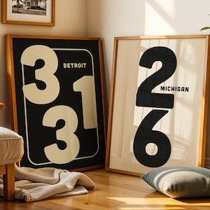 Puede incluir: Dos impresiones enmarcadas con números grandes y nombres de ciudades. Una impresión presenta el número "313" con "DETROIT" arriba. La otra impresión muestra "248" con "MICHIGAN" debajo. Ambas están en marcos de madera, con una paleta de colores neutros.