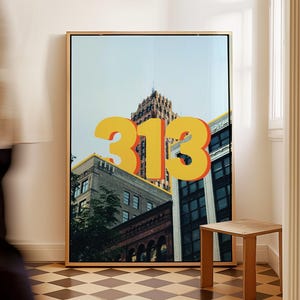 Puede incluir: Una impresión artística enmarcada que presenta un paisaje urbano con el número "313" en letras 3D grandes y amarillas. Los edificios son una mezcla de estilos arquitectónicos, con un fondo de cielo azul claro. Una pequeña mesa auxiliar de madera está en primer plano.