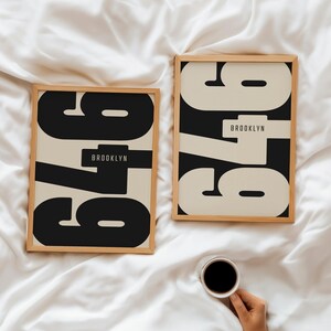 May include: Two framed prints featuring the number "979" in a bold, black font on a cream background. The word "BROOKLYN" is below the numbers. The frames are light wood. A cup of coffee is held in the lower right corner.