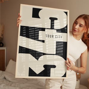 May include: A rectangular framed wall art piece featuring a bold, high-contrast graphic design in black and off-white. The artwork displays large, stylized typography with the text "YOUR CITY" centered in a smaller, clean sans-serif font.