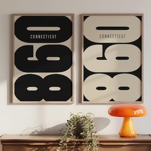 May include: Two framed art prints featuring the word "CONNECTICUT" above the numbers "098" in a bold, monochrome design. The prints are displayed on a wooden dresser with a lamp and plant.