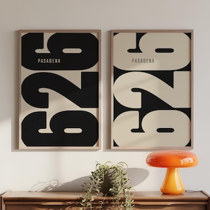 May include: Two framed art prints featuring the word "PASADENA" above large black numbers. The numbers "926" are in a bold font. The background of the prints alternates between black and cream. A wooden dresser holds a plant and an orange mushroom lamp.
