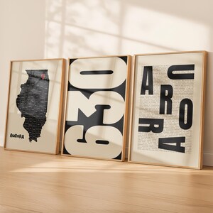 May include: Three framed vertical posters with light beige backgrounds. The left poster shows a black map of Illinois with a red location pin and the text Aurora. The middle poster features large black numbers 630. The right poster displays the word AURORA.