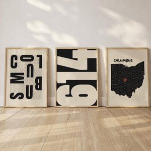Peut inclure: Trois affiches encadrées dans des cadres en bois clair. L'affiche de gauche présente le mot COLUMBUS en grandes lettres noires sur un petit texte. L'affiche centrale affiche le nombre 614 en gras. L'affiche de droite montre une carte noire de l'Ohio avec une épingle rouge.
