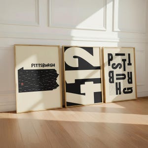 May include: Three framed wall art prints with light wood frames. The first shows a black map of Pennsylvania with the text Pittsburgh and a red location pin. The second features large black and cream numbers 412. The third displays the word Pittsburgh.
