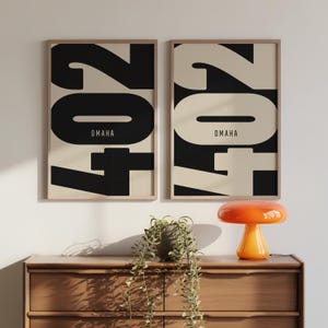 Puede incluir: Dos láminas enmarcadas con el número "204" en negro y la palabra "OMAHA" en una fuente minimalista. El fondo es beige. Una lámpara naranja y una cómoda de madera están en primer plano.