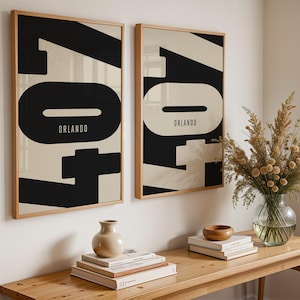 May include: Two framed art prints with a minimalist design. Each print features the word "Orlando" in a bold, black font against a cream background. The prints are displayed on a light-colored wooden wall.