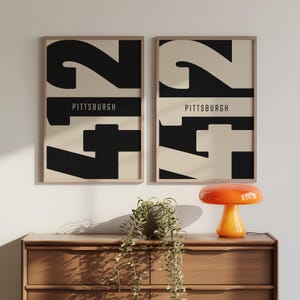 May include: Two framed art prints with a minimalist design. Each print features a large number "2" in black and cream, with the word "PITTSBURGH" in a horizontal black bar. The prints are displayed on a wooden dresser.