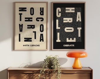 May include: Two framed art prints. One features "NORTH CAROLINA" in bold black letters over a background of smaller text on a cream background. The other print has "CHARLOTTE" in bold white letters on a black background.