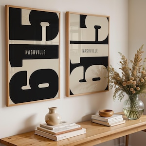May include: Two framed art prints with a mid-century modern design. Each print features the word "Nashville" in white text over a black and beige abstract design. The prints are displayed above a wooden table with decorative items.