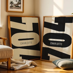 May include: Two framed art prints with a minimalist design. Each print features the word "704" in large black block letters against a cream background, with "CHARLOTTE" printed below. The frames are made of light-colored wood.
