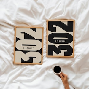 May include: Two framed art prints with the number "302" in black and beige, with "DELAWARE" below. The prints are on a white surface. A hand holds a cup of coffee.
