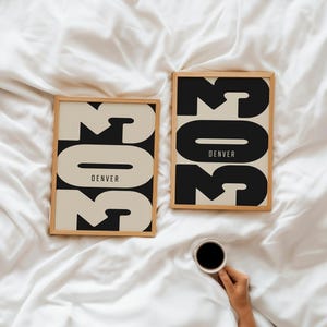 May include: Two framed art prints with the number "303" in a bold, black font, with "DENVER" below. The background of the number is a light beige color. The frames are a light wood color. A hand holds a cup of coffee.