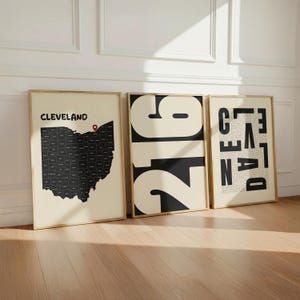 May include: Three framed wall art prints in light wood frames. The left print shows a black map of Ohio with a red location pin and the text CLEVELAND. The center print features the large black numbers 216. The right print displays the word CLEVELAND.