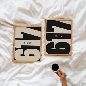Könnte beinhalten: Zwei gerahmte Kunstdrucke mit der Zahl "617" in fetter schwarzer Schrift, darunter "BOSTON". Der Hintergrund der Zahl ist hellbeige. Die Rahmen sind aus hellem Holz. Eine Tasse Kaffee wird in der unteren rechten Ecke gehalten.