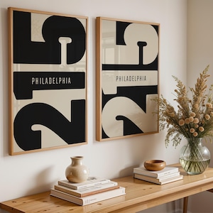 May include: Two framed art prints with a minimalist design. Each print features a large black number 2 and a cream-colored background. The word "PHILADELPHIA" is printed in a black rectangle. The frames are made of light wood.