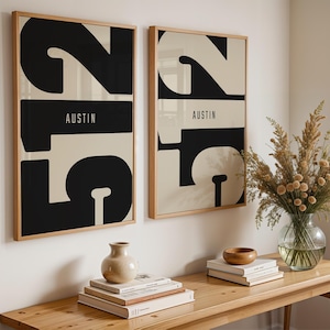 May include: Two framed art prints with a modern design. Each print features a large black number 2 and a stylized number 5 against a cream background, with the word "AUSTIN" in white. The frames are light wood.
