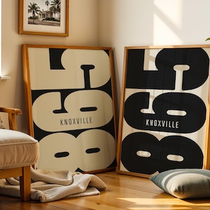 May include: Two framed art prints with a mid-century modern design. Each print features the word "KNOXVILLE" in white text below large black and white abstract numbers. The frames are made of light brown wood. One print is leaning against a wall.