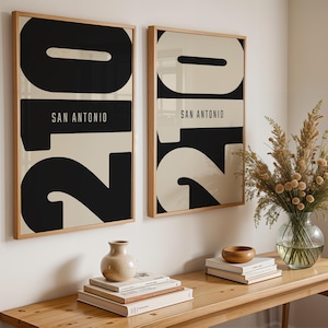 以下が含まれることがあります： ミニマリストデザインの額入りアートプリント2枚。各プリントは、クリーム色の背景に大きな黒い数字「2」と「0」を特徴とし、白いテキストで「SAN ANTONIO」と書かれています。フレームは明るい木材です。プリントは木製のテーブルの上に展示されています。