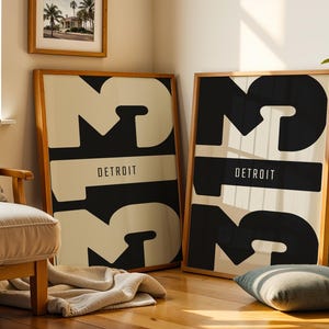 Puede incluir: Dos láminas enmarcadas con un diseño minimalista. Cada lámina presenta un gran número 3 negro con la palabra "DETROIT" en un rectángulo negro. Los marcos son de madera clara y el fondo es blanquecino.
