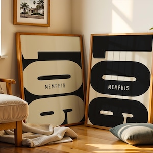 May include: Two framed art prints with a minimalist design. Each print features the word "Memphis" in a black and white color scheme. The artwork is displayed in a room with a wooden floor, a beige sofa, and a framed photograph on the wall.