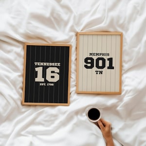 May include: Two framed prints with wooden frames. One features "Tennessee 16 EST. 1796" on a black background with white vertical stripes. The other displays "Memphis 901 TN" on a cream background with vertical stripes. A hand holds a coffee cup.