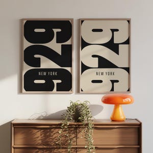 Könnte beinhalten: Zwei gerahmte Kunstdrucke mit der Zahl "626" in fetter, schwarzer Schrift auf cremefarbenem Hintergrund. Der Text "NEW YORK" steht darunter. Die Rahmen sind hellbraun. Eine orangefarbene Pilzlampe steht auf einer Holzkommode.
