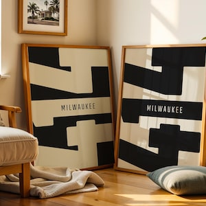 May include: Two framed art prints with a geometric black and cream design, each with the word "MILWAUKEE" in a black rectangle. The frames are made of light-coloured wood. A small framed picture of palm trees hangs above one of the prints.