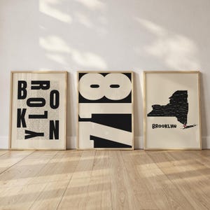 Puede incluir: Tres láminas de arte enmarcadas en madera clara. La lámina de la izquierda muestra la palabra BROOKLYN en letras negras grandes sobre texto pequeño. La central muestra el número 718 en negro negrita. La de la derecha muestra un mapa negro del estado de Nueva York con un alfiler de ubicación rojo sobre la palabra BROOKLYN.