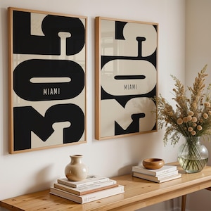 Könnte beinhalten: Zwei gerahmte Kunstdrucke mit minimalistischem Design. Jeder Druck zeigt das Wort "MIAMI" in einer kräftigen Schwarz-Weiß-Grafik. Die Drucke werden an einer weißen Wand über einem Holztisch mit dekorativen Gegenständen ausgestellt.