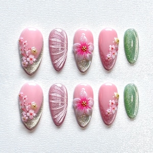 Puede incluir: Un conjunto de ocho uñas artificiales con varios diseños. Las uñas presentan una base rosa con puntas de purpurina plateada, adornadas con flores 3D, purpurina y detalles dorados. Una uña es verde con purpurina.